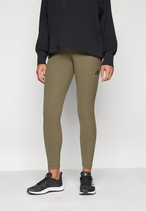 Leggings a coste verde oliva con un design aderente, caratterizzati da un piccolo logo marrone, abbinati a una maglietta nera a maniche lunghe e scarpe da ginnastica nere.
