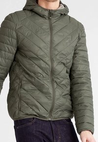 Giacca isolante verde oliva con cappuccio, caratterizzata da un motivo a zigzag trapuntato, zip frontale e polsini elasticizzati. Materiale morbido e leggero.