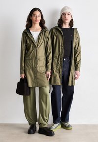 Two individuals wear olive green waterproof jackets with hoods, paired with relaxed green and dark blue pants, and chunky black shoes.