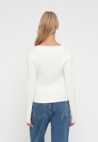 Γυναίκα με μακριά ξανθά μαλλιά, ντυμένη με ένα εφαρμοστό λευκό πλεκτό πουλόβερ και μπλε τζιν σορτς, απεικονίζεται από πίσω σε ένα ενιαίο φόντο.