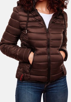 Donna che indossa una giacca trapuntata marrone scuro con zip frontale, cordini del cappuccio e mani nelle tasche con zip, abbinata a jeans blu.