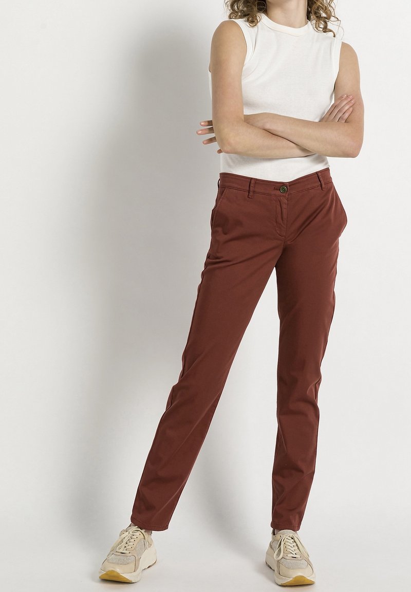 Slim-fit kalhoty v rezavé barvě s hladkou texturou, s předním zapínáním na knoflík a bočními kapsami. Doplněno bílým topem bez rukávů.