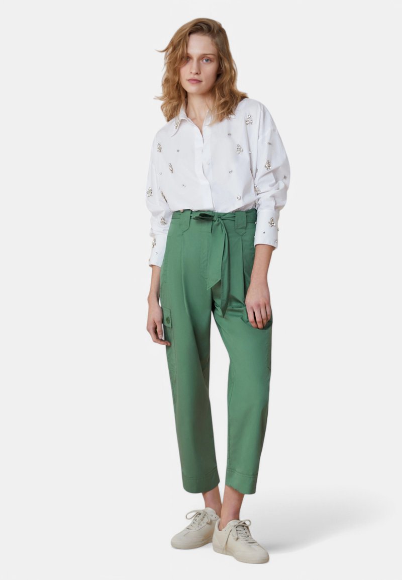 Camicia bianca con ricami floreali e pantaloni verdi con vita alta, dotati di cintura, tasche laterali e orli a risvolto. Sneakers bianche casual.