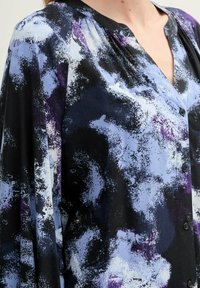 Blusa in tessuto leggero con una stampa astratta in nero, blu e viola. Presenta una chiusura frontale con bottoni e un dettaglio con scollo plissettato.