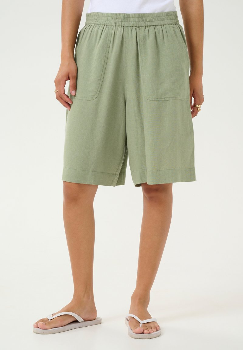 Shorts vert clair, jusqu'aux genoux, avec une taille élastique, deux poches latérales et un tissu texturé. Portés avec des tongs simples.