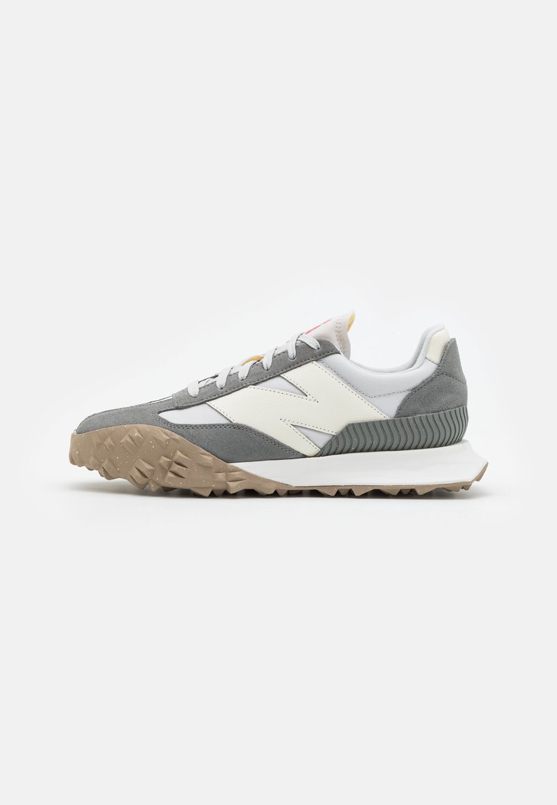 Chaussure de sport profil bas en suède gris, dessus en cuir blanc, semelle beige texturée et lacets blancs, vue de côté sur fond blanc.