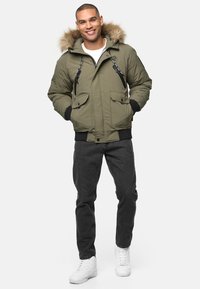 Olivgrüne Pufferjacke mit einem gefütterten Kapuzenrand, Reißverschluss, zwei vorderen Taschen und schwarzen Rippbündchen, kombiniert mit dunklen Jeans und weißen Sneakers.