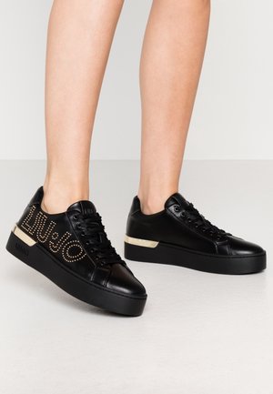 Beine mit schwarzen Plateau-Sneakern, mit goldbesetztem "Liu Jo"-Logo an der Seite und goldenem Akzent an der Ferse, stehend auf hellem Boden.