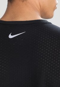 Schwarzes Sportshirt aus einem Netzgewebe, mit rundem Halsausschnitt und einem kleinen weißen Nike-Logo auf der Rückseite, das unterhalb des Nackens platziert ist.