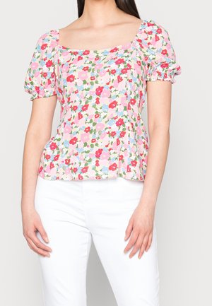 Femme portant un chemisier à imprimé floral avec des manches bouffantes et un col carré, associé à un pantalon blanc, debout devant un fond clair.