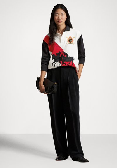 Polo blanc avec accents rouges et noirs, présentant un graphique de cavalier ; associé à un pantalon noir taille haute et une pochette noire.