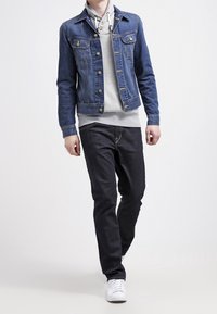 Chaqueta de denim en azul oscuro, con cierre de botones a presión y bolsillos en el pecho, sobre una sudadera con capucha gris clara, combinada con jeans oscuros y zapatillas blancas.
