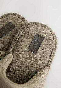 Paire de chaussons en tissu beige texturé avec des patchs noirs rectangulaires portant l'étiquette "NEXT" sur la semelle intérieure sur fond blanc.