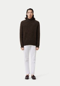 Brauner, grob gestrickter Pullover mit hohem Kragen, kombiniert mit weißen Jeans und dunklen Slippern. Der Pullover hat ein strukturiertes Muster.