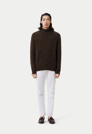Un pull en tricot marron et épais avec un col haut, associé à un jean blanc et des mocassins foncés. Le pull présente un motif texturé.