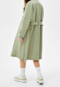 Długi, jasnobrązowy płaszcz z paskiem, kieszeniami bocznymi i wentylacją z tyłu. Nosi się go na białych skarpetach i beżowych sneakersach z akcentami.