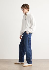 Pullover blanc en polaire avec fermeture éclair quart, pantalon en denim bleu à jambes larges, et baskets blanches avec une semelle en caoutchouc. Le modèle pose de manière décontractée contre un fond neutre.
