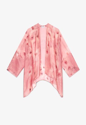 Joven mujer con cabello ondulado lleva pantalones anchos rosa con efecto tie-dye y un kimono transparente a juego sobre una camiseta blanca, de pie sobre un suelo de hormigón.