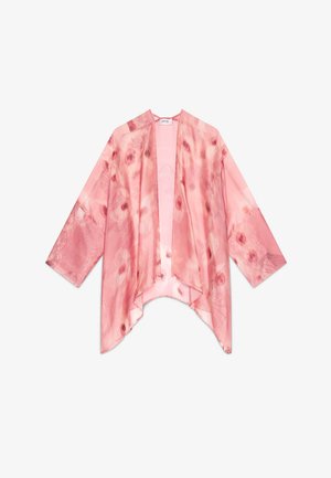 Joven mujer con cabello ondulado lleva pantalones anchos rosa con efecto tie-dye y un kimono transparente a juego sobre una camiseta blanca, de pie sobre un suelo de hormigón.