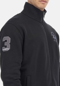Fekete, cipzáros pulóver magas gallérral, a jobb ujján szürke "3"-as szám és a mellkasán logó található. Puha, texturált anyag.