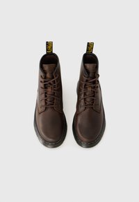 Les bottes en cuir marron montantes présentent un bout rond, un laçage à l'avant et une tirante avec le logo jaune. Semelle en caoutchouc noir texturé avec un détail de coutures.