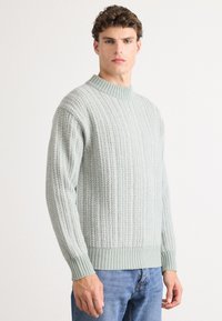 Man wearing light gray ribbed knit sweater and blue jeans standing against white background, facing forward with neutral expression.