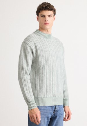 Man wearing light gray ribbed knit sweater and blue jeans standing against white background, facing forward with neutral expression.