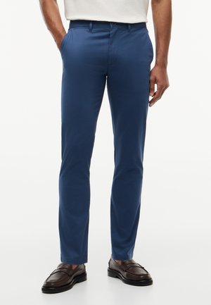 Άνδρας που φοράει εφαρμοστά μπλε παντελόνια, λευκό πλεκτό πουλόβερ και σκούρα καφέ δερμάτινα μοκασίνια, στέκεται με το ένα χέρι στην τσέπη.