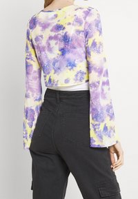 Długi rękaw crop top z przezroczystego materiału w fioletowo-żółty wzór tie-dye. Czarny wysoki pas spodni z kieszeniami i delikatną teksturą.