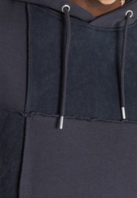 Felpa con cappuccio patchwork blu navy in tessuto testurizzato, con occhielli e cordoncini argentati. Le cuciture in vista mettono in risalto il design a strati.