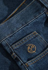 Jeans de mezclilla azul oscuro con costuras en color beige. Un pequeño logo está bordado en el bolsillo trasero, con una superficie de tela suave y texturizada.