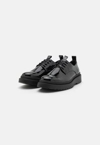 Zapatos de charol negro con cordones, de puntera redonda y suela de goma negra gruesa; poseen una superficie lisa y brillante, y costuras decorativas.