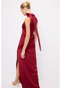 Rød satin kjole med høj krave, åben ryg og lang sløjfe. Har en side-split og glat tekstur. Elegant silhuet.