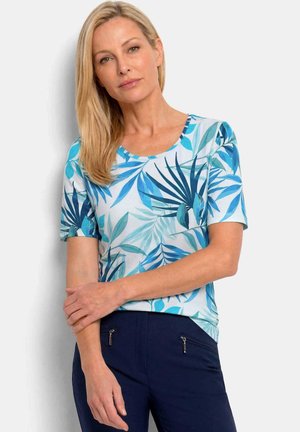 Femme blonde portant une chemise à manches courtes bleu et turquoise à motif feuilles et un pantalon bleu marine foncé, debout les bras croisés.