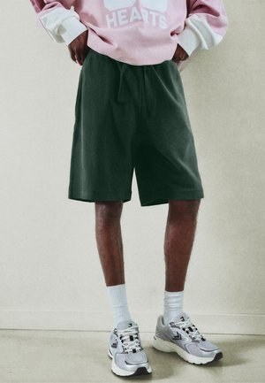 Osoba nosiaca tmavozelené šortky po kolená, biele ponožky, sivé bežecké topánky a ružovú mikinu s bielymi manžetami, stojaca vo vnútri.