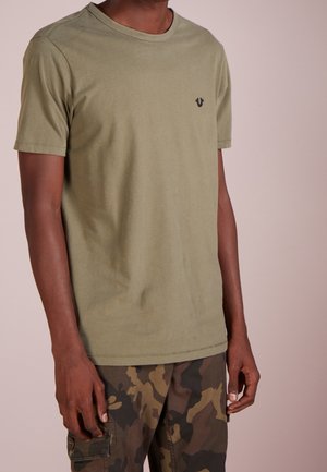 Person iført en olivengrøn kortærmet t-shirt og brun-grøn camouflage cargobukser, stående mod en ensfarvet baggrund.