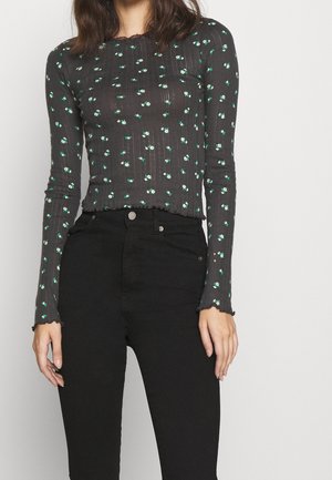 Frau trägt ein eng anliegendes schwarzes Langarmshirt mit kleinem grünem Blumenmuster und eine hochgeschnittene schwarze Hose, vom Hals bis zu den Hüften gezeigt.