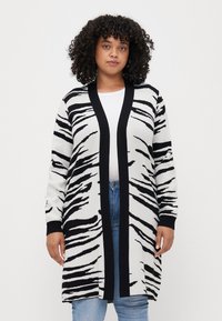 White and black zebra-striped long cardigan with a black open front, long sleeves, and ribbed cuffs, worn over a white shirt and jeans.