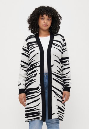 Hvit og svart zebra-striped lang cardigan med åpen front i svart, lange ermer og ribbede mansjetter, fås over en hvit skjorte og jeans.