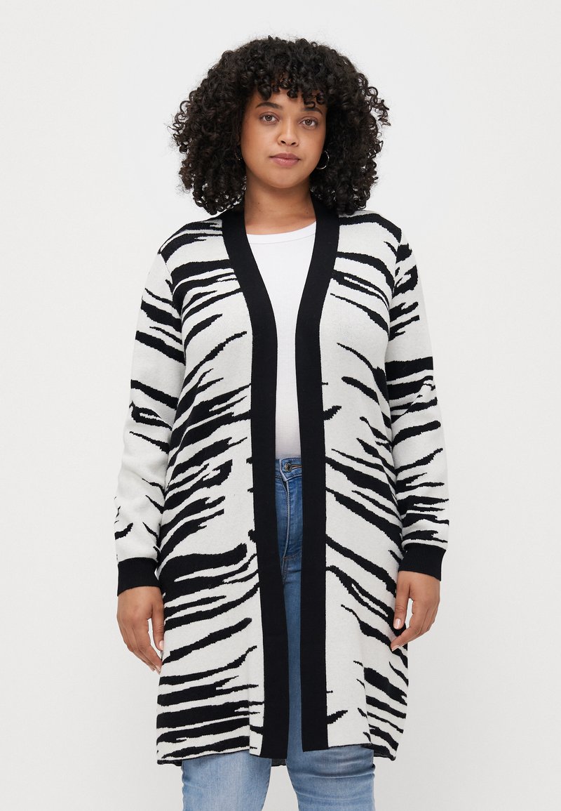 White and black zebra-striped long cardigan with a black open front, long sleeves, and ribbed cuffs, worn over a white shirt and jeans.