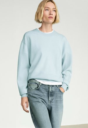 Donna bionda con un maglione azzurro chiaro e jeans blu slavati, con una mano in tasca, in piedi davanti a uno sfondo chiaro e uniforme.