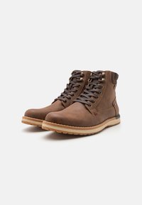 Botas de tobillo de cuero marrón con punta redonda, cierre de cordones, suela beige en contraste y parte superior texturizada. Presenta ojales de metal y un pequeño logo.