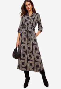 Rochie cămașă cu model negru și crem, cu mâneci lungi, talie accentuată cu curea, buzunare laterale și fustă fluidă. Asortată cu o geantă texturată neagră.