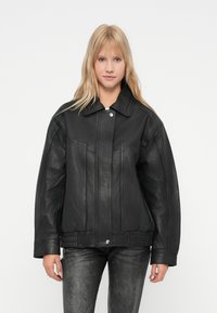 Jonge vrouw met lang blond haar, gekleed in een zwart leren jack en donker gewassen jeans, staand tegen een effen lichte achtergrond.