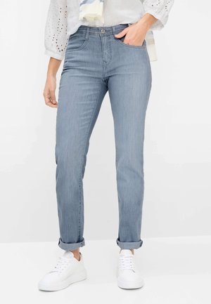 Persona che indossa jeans blu chiaro risvoltati, sneakers bianche, camicetta bianca con maniche a occhiello e una leggera sciarpa, in piedi con una mano in tasca.