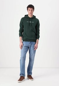 Dunkelgrüner Kapuzenpullover mit einer Fronttasche und Kordelzug. Kombiniert mit hellblauen Jeans und braunen Sneakern. Text auf dem Kapuzenpullover in Weiß.