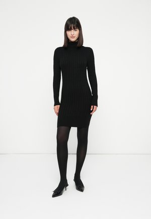 Vrouw die staat in een zwarte kabelgebreide coltrui jurk, zwarte panty’s en zwarte puntige hakken tegen een witte achtergrond.