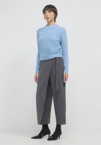 Femme debout portant un pull léger bleu, un pantalon large gris avec un lien sur le côté, des collants noirs et des chaussures à talons bas noires sur fond blanc.
