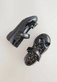 Chaussures noires en cuir verni avec un détail en nœud, bride réglable et semelle intérieure à motifs de nœuds. Semelle en caoutchouc pour une meilleure adhérence.