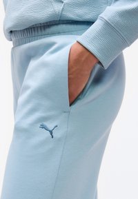 Το χέρι ενός ατόμου στην τσέπη ανοιχτόχρωμων γαλάζιων παντελονιών φόρμας Puma, φορώντας ένα ίδιο υφασμάτινο μακρυμάνικο πάνω μέρος.